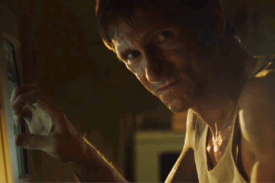 Horror icon Bill Oberst Jr. in the Emmy Award-winning "Take This Lollipop." Bill stares at the viewer while lightly touching a desktop computer monitor in a dark basement. He is sweaty and dressed in a grimy wifebeater. 